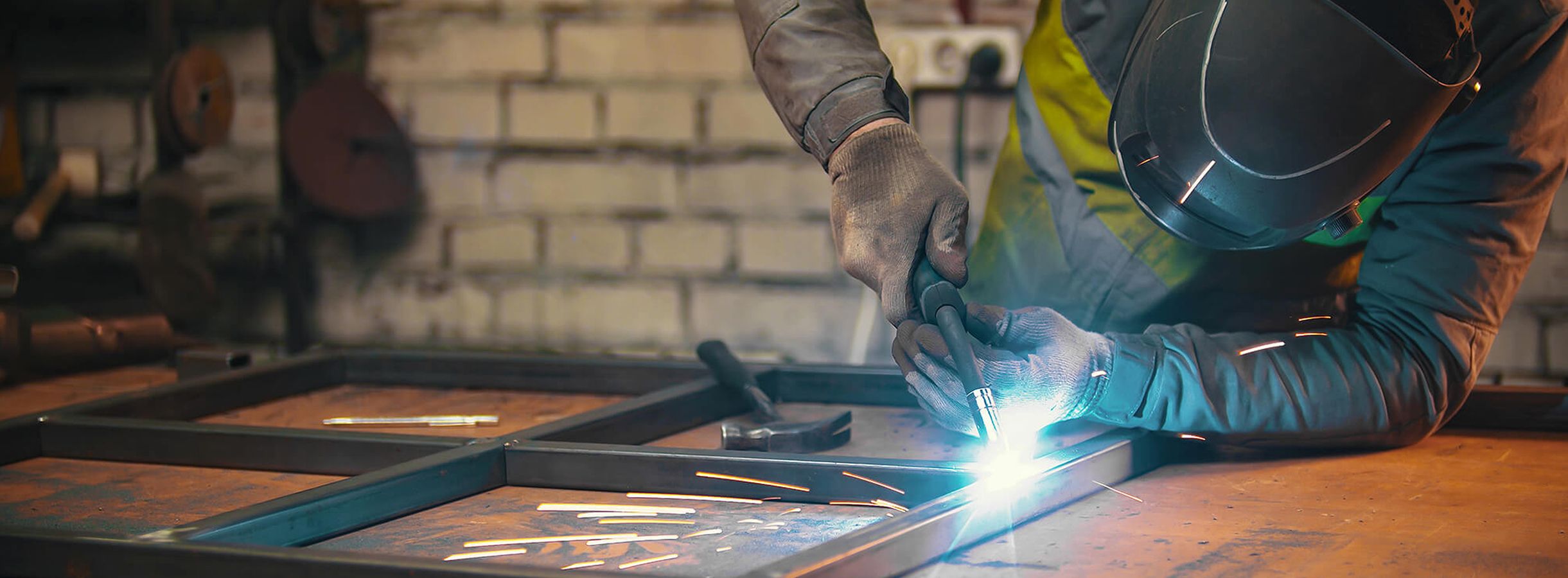 Person mit Schutzhelm und Handschuhen schweißt an einem Metallrahmen auf einem Arbeitstisch